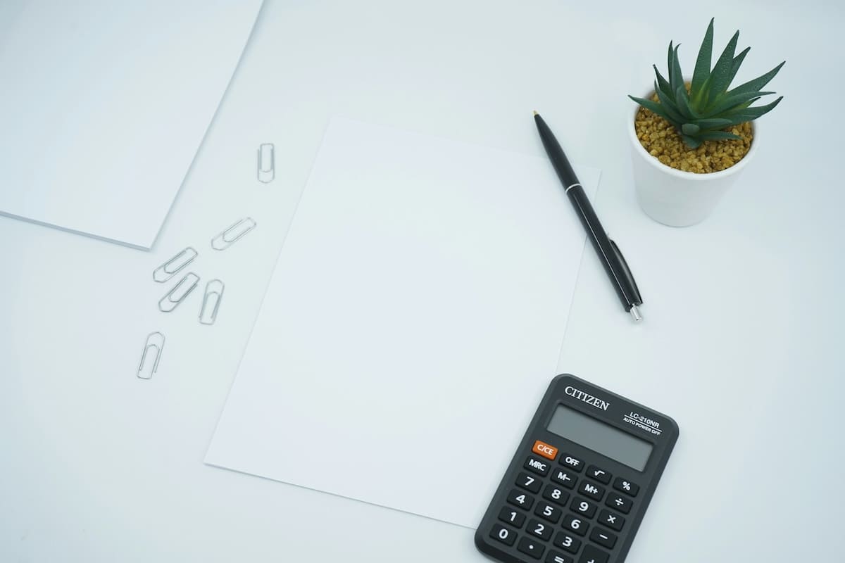白い机に置かれた電卓と紙、ペン、観葉植物